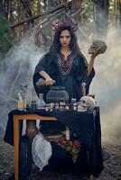 Witch in black, long dress, with red crown in her hair. Posing in pine forest, holding skull, making potion. Spells and witchcraft. Close-up, smoke. photo