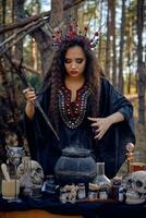 Witch in black, long dress, with red crown in her long hair. Posing in pine forest. Making a magic potion. Spells and witchcraft. Close-up, smoke. photo