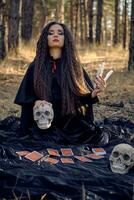 Witch in black dress with cape and hood. Posing in pine forest. Sitting on dark blanket whith fortune-telling cards and skulls on it. Full length. photo