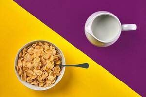 Bowl with corn flakes, jug of milk and spoon on purple and yellow background, top view photo