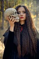 Witch in black, long dress with cape and hood. Posing in smoke pine forest. Holding a skull. Spells, magic and witchcraft. Close-up. photo