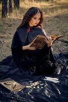 Witch in black dress with cape and hood. Posing in pine forest. Sitting on dark blanket whith books, candles and runes on it. Full length. photo