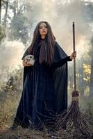 Witch in black, long dress with cape and hood. Posing in smoke pine forest. Holding a skull and a broom. Spells, magic and witchcraft. Full length. photo