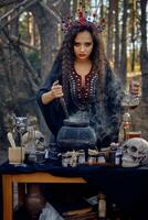 Witch in black, long dress, with red crown in her long hair. Posing in pine forest. Making a magic potion. Spells and witchcraft. Close-up, smoke. photo