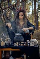 Witch in black, long dress, with red crown in her long hair. Posing in pine forest. Making a magic potion. Spells and witchcraft. Close-up, smoke. photo