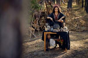 Beautiful witch in black, long dress, with red crown in her long hair. Posing in pine forest. Spells, magic and witchcraft. Full length portrait. photo