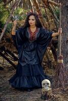 Witch in black, long dress, with red crown in her long hair. Posing with broom and skull in pine forest. Spells, magic and witchcraft. Full length. photo