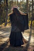 Witch in black, long dress, with red crown in her long hair under a black veil. Posing in pine forest. Spells, magic and witchcraft. Full length. photo