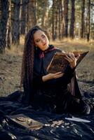 Witch in black dress with cape and hood. Posing in pine forest. Sitting on dark blanket whith books, candles and runes on it. Full length. photo