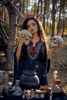 Witch in black, long dress, with red crown in her long, curly hair. Posing in pine forest. Holding skulls. Spells, magic and witchcraft. Close-up. photo