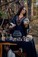 Witch in black, long dress, with red crown in her hair. Posing in pine forest, holding skull, making potion. Spells and witchcraft. Close-up. photo