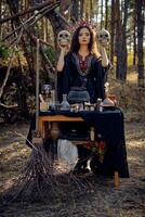 Witch in black, long dress, with red crown in her long hair. Posing in pine forest. Holding skulls. Spells, magic and witchcraft. Full length. photo