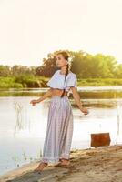 On the edge of the shore near the river stands a girl in a white dress. photo