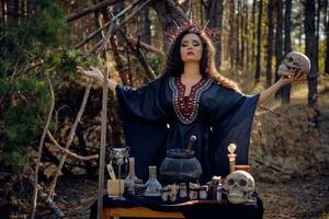 Witch in black, long dress, with red crown in her long, curly hair. Posing in pine forest. Holding skull. Spells, magic and witchcraft. Close-up. photo