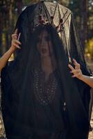 Witch in black, long dress, with red crown in her long, curly hair under a black veil. Posing in pine forest. Spells, magic and witchcraft. Close-up. photo