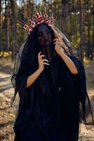 bruja en negro, largo vestido, con rojo corona en su largo, Rizado pelo debajo un negro velo. posando en pino bosque. hechizos, magia y brujería. de cerca. foto