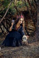 Witch in black, long dress, with red crown in her long hair. Posing with broom and skull in pine forest. Spells, magic and witchcraft. Full length. photo