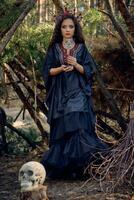 Witch in black, long dress, with red crown in her long hair. Posing with broom and skull in pine forest. Spells, magic and witchcraft. Full length. photo