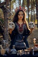 Witch in black, long dress, with red crown in her long, curly hair. Posing in pine forest. Holding skulls. Spells, magic and witchcraft. Close-up. photo