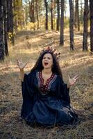 Beautiful witch in black, long dress, with red crown in her long hair. Posing sitting in pine forest. Spells, magic and witchcraft. Full length. photo