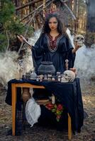 Witch in black, long dress, with red crown in hair. Posing in pine forest, holding skull, making potion. Spells and witchcraft. Full length, smoke. photo