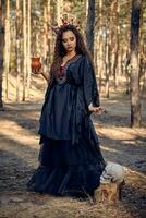 Witch in black, long dress, with red crown in hair. Giving water to skull from pot while posing in pine forest. Spells, witchcraft. Full length. photo