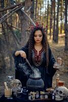 Witch in black, long dress, with red crown in her long hair. Posing in pine forest. Making a magic potion. Spells and witchcraft. Close-up, smoke. photo