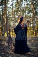 Witch in black, long dress, with red crown in her long hair. Posing with broom and skull in pine forest. Spells, magic and witchcraft. Full length. photo