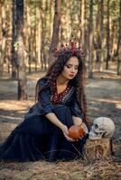 Witch in black, long dress, with red crown in hair. Giving water to skull from pot while sitting in pine forest. Spells, witchcraft. Full length. photo