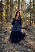 Witch in black, long dress, with red crown in her long hair. Posing sitting on broom in pine forest. Spells, magic and witchcraft. Full length. photo