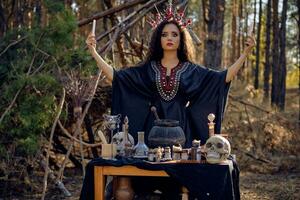 hermosa bruja en negro, largo vestido, con rojo corona en su largo, Rizado cabello. posando en pino bosque. hechizos, magia y brujería. de cerca. foto