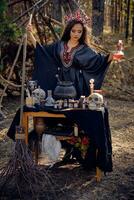 Beautiful witch in black, long dress, with red crown in her long hair. Posing in pine forest. Spells, magic and witchcraft. Full length portrait. photo