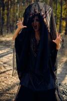 Witch in black, long dress, with red crown in her long, curly hair under a black veil. Posing in pine forest. Spells, magic and witchcraft. Close-up. photo