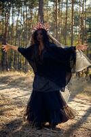 Witch in black, long dress, with red crown in her long hair under a black veil. Posing in pine forest. Spells, magic and witchcraft. Full length. photo