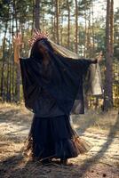 Witch in black, long dress, with red crown in her long hair under a black veil. Posing in pine forest. Spells, magic and witchcraft. Full length. photo