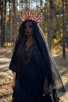 Witch in black, long dress, with red crown in her long, curly hair under a black veil. Posing in pine forest. Spells, magic and witchcraft. Close-up. photo