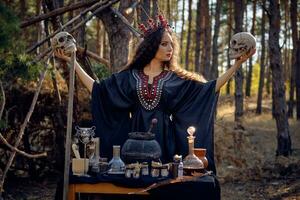 Witch in black, long dress, with red crown in her long, curly hair. Posing in pine forest. Holding skulls. Spells, magic and witchcraft. Close-up. photo