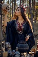 Witch in black, long dress, with red crown in her long, curly hair. Posing in pine forest. Holding skull. Spells, magic and witchcraft. Close-up. photo