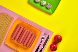 Sausages with tomatoes, spaghetti and garlic on a yellow and pink minimal background. Flat lay. Top view. photo