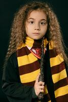 Portrait of a little witch girl with long brown hair dressed in dark coat, holding magic wand in her hand, posing on black studio background. photo