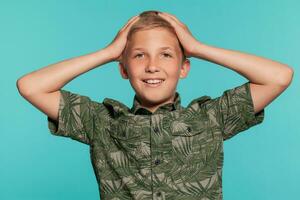 Close-up portrait of a blonde teenage boy in a green shirt with palm print posing against a blue studio background. Concept of sincere emotions. photo