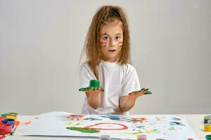 pequeño niña en blanco camiseta sentado a mesa con qué hombre y pinturas en él, posando con pintado cara y manos. aislado en blanco. medio de cerca. foto