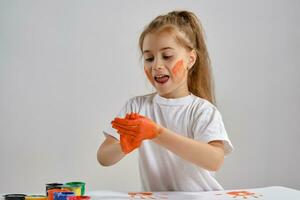 Little girl in white t-shirt sitting at table with whatman and colorful paints on it, painting her hands. Isolated on white. Medium close-up. photo