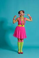 encantador niña con un multicolor trenzas peinado y brillante constituir, posando en estudio en contra un azul fondo, participación un pirulí en su mano. foto