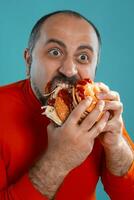 Close-up portrait of a middle-aged man with beard, dressed in a red turtleneck, posing with burgers against a blue background. Fast food. photo