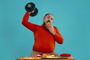 Close-up portrait of a middle-aged man with beard, dressed in a red turtleneck, posing with burgers and french fries. Blue background. Fast food. photo