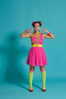 encantador niña con un multicolor trenzas peinado y brillante constituir, posando en estudio en contra un azul fondo, participación un pirulí en su mano. foto