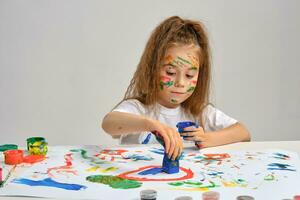 Little girl in white t-shirt sitting at table with whatman and paints, drawing on it, posing with painted face, hands. Isolated on white. Close-up. photo