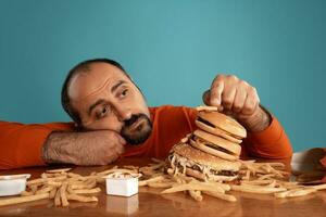 de cerca retrato de un de edad mediana hombre con barba, vestido en un rojo suéter tipo con cuello de tortuga, posando con hamburguesas y francés papas fritas azul antecedentes. rápido alimento. foto