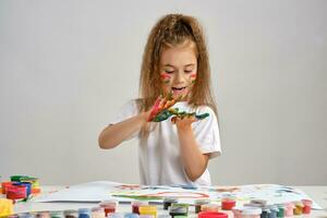 Little girl in white t-shirt sitting at table with whatman and paints on it, posing with painted face and hands. Isolated on white. Medium close-up. photo